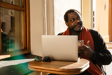 Black bearded man using mobile phone and laptop while sitting in cafe