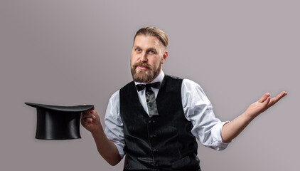 Portrait of bearded mature magician in suit holding black top hat while posing over grey...
