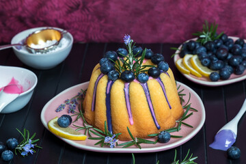 Guglhupf mit Zuckerguß, Blaubeeren, Rosmarin, Zitrone und Puderzucker auf rosafarbenen Teller