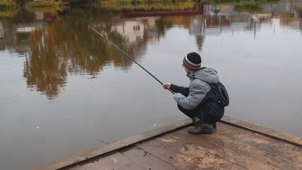 A teenage Caucasian boy is fishing on a calm autumn day on the lake squatting