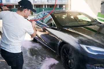 Male driver in white shirt using high pressure water sprayer while washing modern electric car on...