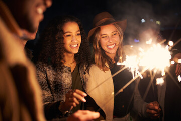 Multi-cultural best friends at a rooftop party smiling while lighting sparklers. Night time,...