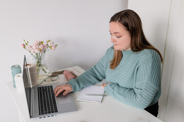 Young stylish woman using laptop at home, looking at screen, chatting, reading or writing email. Female hobby. Working on research project online concept. Handmade macrame shop