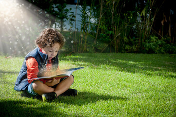 Criança sentada no gramado de colete azul e bermuda lendo livro, com raios de sol vindo do...