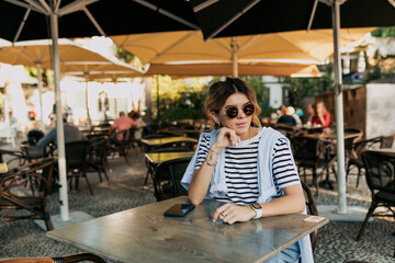 Obraz na płótnie Canvas Stylish lovely woman with collected hair in sunglasses wearing striped t-shirt and blue shirt sitting on summer terrace and looking away. Recreation concept. Weekend time