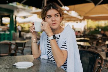 Adorable lovely woman with collected hair wearing striped t-shirt and blue shirt sitting on open air cafe and drinking coffee in warm sunny day