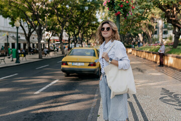 Outdoor photo of caucasian girl with snow-white smile looks into camera. Against background of city with palms and city cabs, short-haired beauty in sunglasses with eco bag