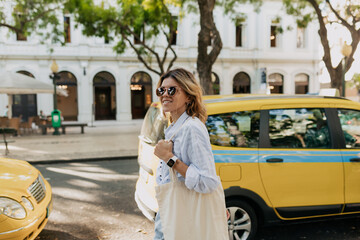 Obraz premium Happy excited girl with wavy short light hairstyle dressed blue shirt walking with eco bag in sunny old city. Outdoor portrait of tourist happy woman exploring new place