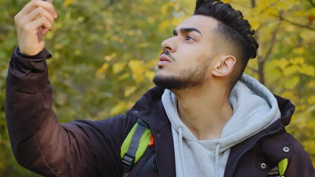 Young man standing outdoors lagged behind tourist group lost in wood traveling with backpack answering mobile call no connection frustrated looking at smartphone screen worried about internet problem