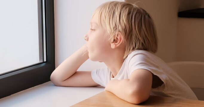 Bored blond boy sits at table and looks sadly out the window. Child boy is waiting for something near the window.