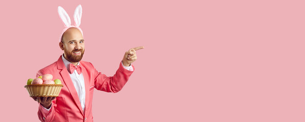 Look here. Happy handsome bearded man in suit, bow tie and cute bunny ears standing on pink background, holding wicker bowl of Easter eggs and pointing finger to side at blank copyspace background