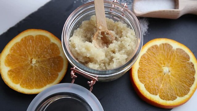 Homemade sugar body scrub in glass jar, decorated with fresh orange slices and wooden spoon with sugar powder on black stone cutting board. Body skin care concept.
