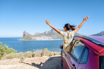 woman outside car window road trip with hands up, car at Chapmans Peak Drive in Cape Town South Africa looking out over the ocean.  - obrazy, fototapety, plakaty