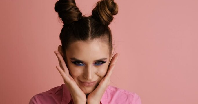 Cheerful young girl with two buns laughs and dances, makes surprised face. Close up video models in studio on pink background.
