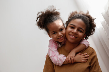 Younger and older sister spending time together at home. Two black girls of different age hugging...