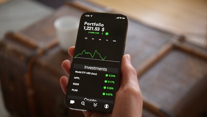 Hand holding Phone with Trading App Mock-up at home in the living room, watching Stock Market and Investments rising in Dollar. Trend of young people investing in stocks.