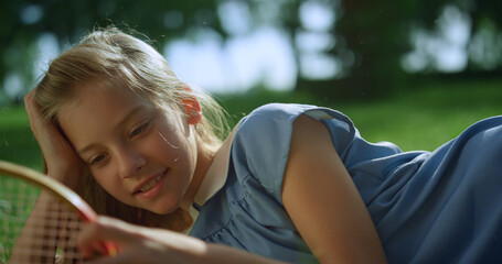 Smiling girl lying on green lawn alone. Blond kid touch racket net closeup
