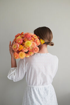 Conceptual portrait of young woman wearing white cotton dress covering her face with lush ranunculus. Female hiding behind beautiful bouquet of bright flowers. Close up, copy space, white background