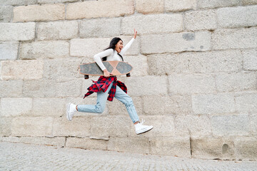 young latin hipster girl jumping around happily carrying a skateboard