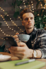 Low angle of serious male entrepreneur with smartphone drinking coffee during distant work in cafe 