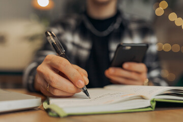 Crop male freelancer taking notes in notebook and browsing smartphone while working online in cafe 