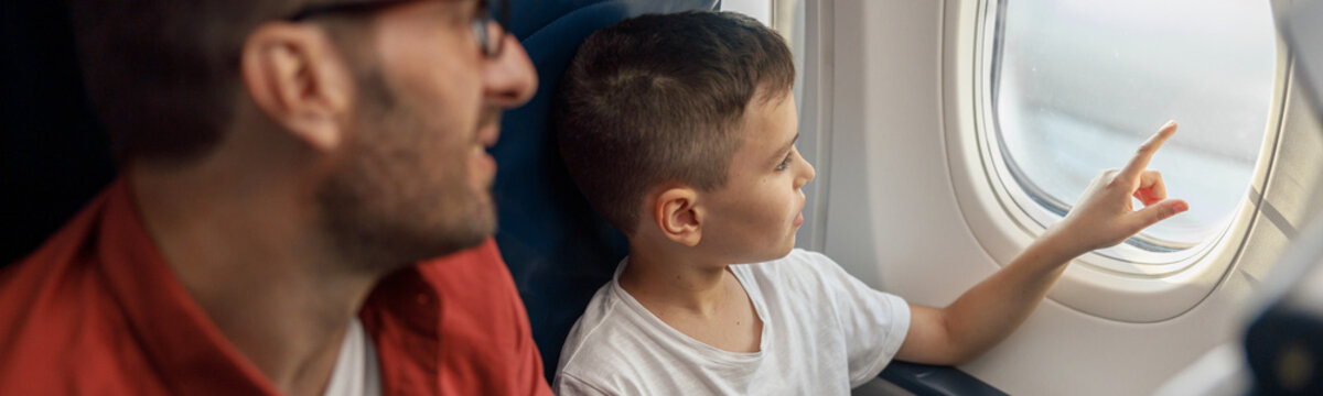 Curious Little Boy Looking Out The Window While Traveling By Plane Together With His Father. Family, Transportation, Vacation Concept