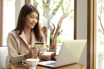 Asian woman feeling happy using credit card shopping online with computer laptop and drinking coffee in coffee shop. Female consumer buying things on internet, using contactless payment.