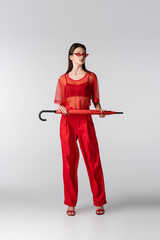 full length of young woman in red outfit and sunglasses posing with umbrella on grey.