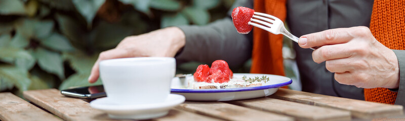 Mature woman eats delicious toast with cream and strawberries and takes mobile phone near cup at...