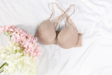 Sexy women's underwear and flowers on the bed. Beige color bra, lace lingerie on a white bedsheets background. Beauty blog concept. Top view, flat lay