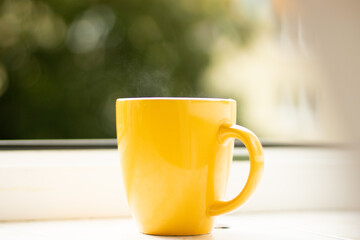 Morning tea on the windowsill from which steam comes out. Yellow cup with a drink near the open...