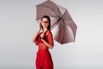 young woman in red outfit adjusting sunglasses and standing under umbrella isolated on grey.
