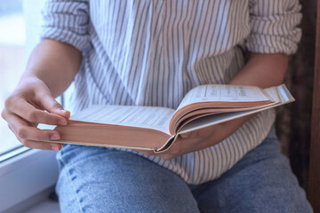 A young female student with a book sitting on window sill, studying.