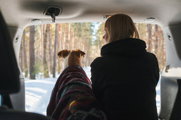 Winter car camping with pets scene: woman sits next to a dog in SUV trunk. Enjoying the early spring and sun in the forest, exploring nature with pets
