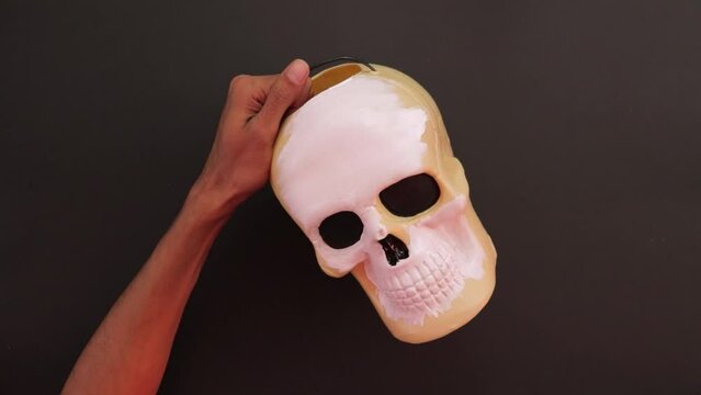Painting a candy skull bucket with white paint. Mexican decorations. Brown skin hands. Black background. 