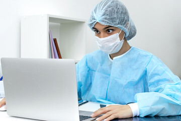 Female doctor wearing face mask and surgical overalls is sitting in front of a laptop screen in a medical office.Healthcare and medical concept.