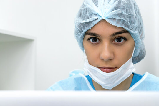 Female Doctor Wearing Face Mask And Surgical Overalls Is Sitting In Front Of A Laptop Screen In A Medical Office.Healthcare And Medical Concept.