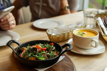 a frying pan with fried meat and vegetables in the foreground stands on a wooden table