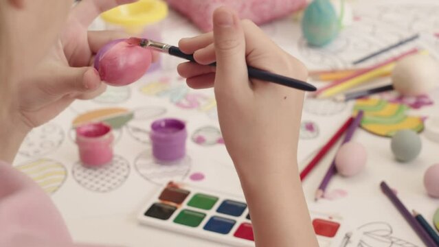 Slowmo closeup of little girl coloring Easter egg in pink with brush sitting at table with paints, coloring pages and other Easter decorations