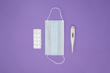 Top view, medical accessories on purple background. Mask, thermometer, medicines, pills. Flat lay
