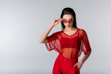 young model in red trendy outfit adjusting sunglasses while posing with hand in pocket isolated on...