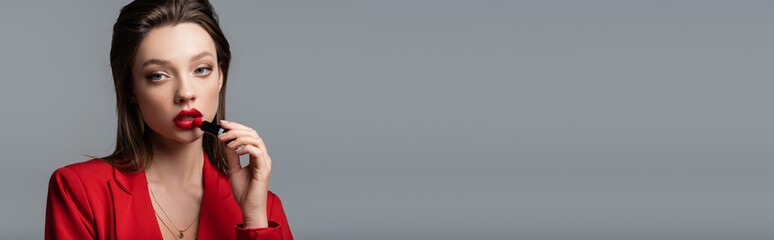 young woman in red blazer applying lipstick isolated on grey, banner.