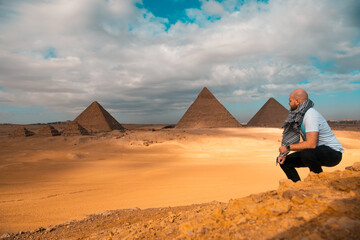Man sitting on the sandy desert dunes posing in front of the great pyramids of giza. Traveling...