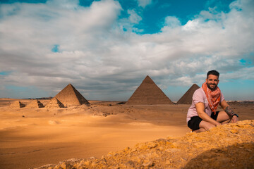 Man sitting on the sandy desert dunes posing in front of the great pyramids of giza. Traveling egypt in winter time, tourists posing for a picture