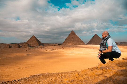 Man sitting on the sandy desert dunes posing in front of the great pyramids of giza. Traveling egypt in winter time, tourists posing for a picture, looking at the pyramids