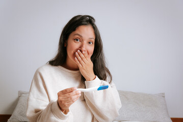 young woman looking in camera holding pregnancy test with surprised face. hispanic woman in her thirties smiling discovering a baby coming and new maternity
