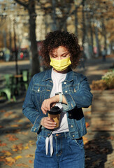 Beautiful curly haired woman in protective mask holding a cup of coffee near the outdoor cafes.
