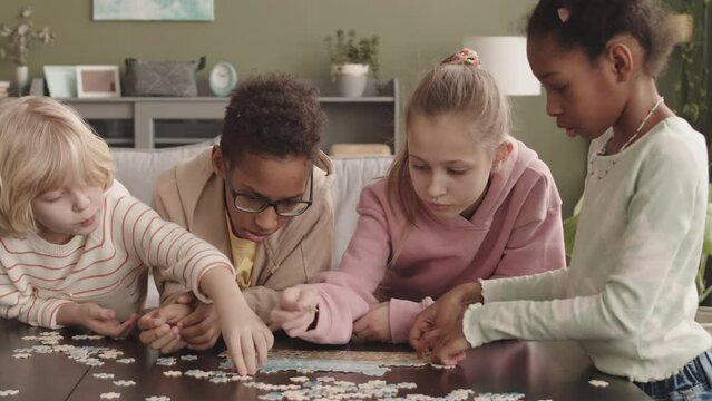 Waist up slowmo of four multiethnic elementary age kids solving jigsaw puzzle together on table at home