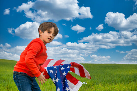 Child With The Flag Of The United States, He Is On A Green Meadow With A Dramatic Sky, He Is Wearing A Red t-shirt And Jeans. Image With Copy Space. Nature And People