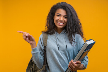 Studio image of cute smiling casually dressed student girl looking directly to the camera and poiting with her index finger to the left side, isolated over yellow background